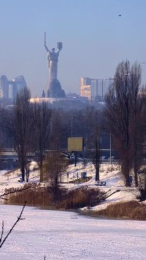 Görkemli Ukrayna Ana anıtı soğuk ve soğuk bir kış gününde gökyüzünde yükseliyor. Bir kuş donmuş, karla kaplı bir parkın üzerinde uçuyor ve arka planda sisli bir gökyüzü var.