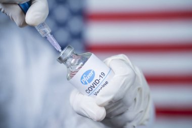 Maski, India - Nov 12,2020 : Doctor holding Pfizer Biontech vaccine and syringe to protect against coronavirus COVID-19 disease with US flag as background clipart