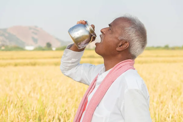 Thirsty Indian farmer drinking water from steel tumbler or Chambu on hot sunny day at agriculture field.