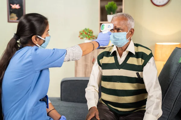 Врач или медсестра проверяют температуру пожилого больного человека с помощью инфракрасного термометра в больнице, в то время как оба носят маску для лица из-за коронавируса ковид-19 меры безопасности — стоковое фото