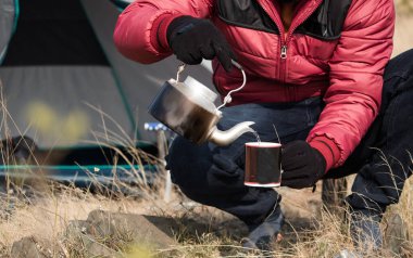 Tanımlanamayan Gezgin elleri çay fincanının içine sıcak su döküyor. Kamp sırasında kamp ateşi kullanarak çaydanlığı ısıtan yürüyüşçü kavramı.