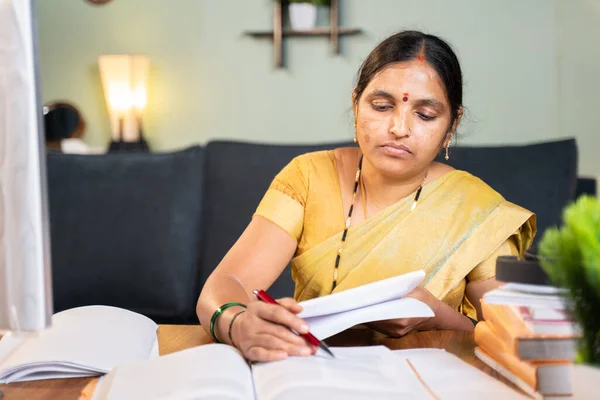 Indian woman professor correcting answer sheets of books by referring books at home at working desk