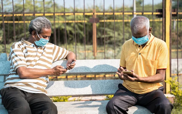 Two elderly people with medical face mask busy using mobile phone while sitting at park while maintaining social distance - concept seniors using smartphone, technology, social media and apps.