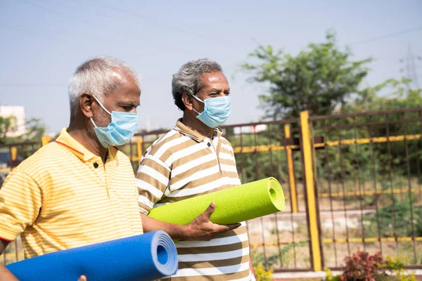 Deux hommes âgés avec masque médical tenant tapis de yoga viennent au parc pour faire de l'exercice le matin concept de nouveaux soins de santé normaux et de remise en forme pendant la pandémie de coronavirus covid-19. — Photo