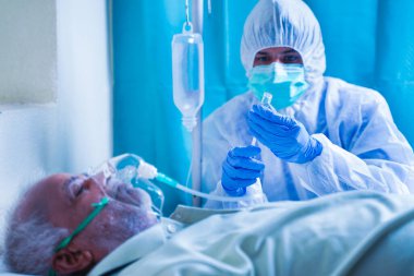 Doctor with potective suit preparing injection syringe while patient breathing on ventilator oxygen mask due to coronavirus covid-19 viral infection at hospital