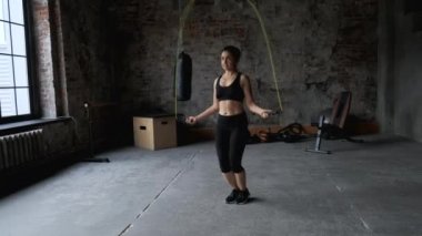 Koyu renk spor salonunda ip atlayan genç bir kadının portresi. Kadın sporcu içeride antrenman yapıyor. 