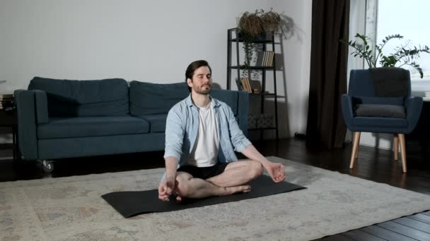 年轻健康的男人做瑜伽 荷花的姿势 在他的公寓地板上 他紧闭双眼坐在黑色的垫子上 — 图库视频影像