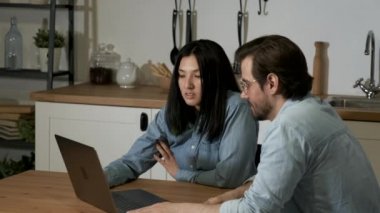 Close-Up of a Young European Man Talking to an Asian Woman, Discussing a New Project Business Smartphone App, Working Online at Home in the Kitchen