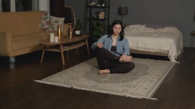 Relaxed young Asian woman in sportswear is meditating in lotus position sitting on floor with closed eyes at home enjoying meditation with hands in namaste.