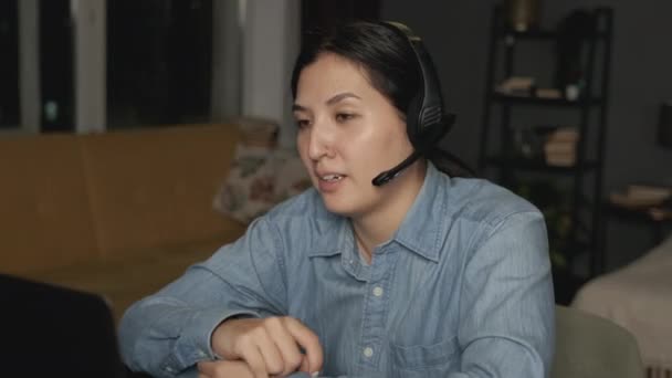 サイドビューマイクを持つヘッドフォンの若いアジアの女性のクローズアップ自宅の暗い部屋でラップトップを使用して クライアントオンラインビデオ会議と通信する — ストック動画