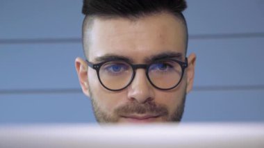 Dizüstü bilgisayarın önünde gözlüklü, gözlüklü genç Avrupalı bir işadamı gözlüklü bir şekilde yansıyor. Serbest çalışan genç bir erkek.