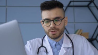 Erkek doktorun tıbbi belgelere baktığı ve ofiste otururken dizüstü bilgisayarda daktilo yazdığı görüntüleri kapatın. Gözlüklü genç adam ve iş yerindeki beyaz profesyonel soygunun görüntüsü.