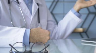 Doktorun ellerinin bir şeyi açıklarken ve gösterirken yakın plan çekimleri masanın üzerinde yansıması olan gözlükler var.