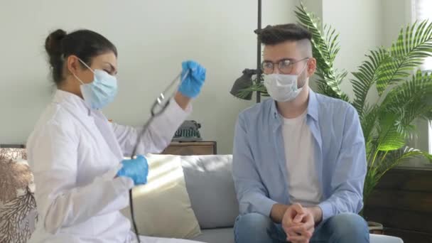 Aマスクの若い女性医師は 流行性ウイルス中の男性患者を聞きます 明るい居心地の良い部屋に家にあります — ストック動画