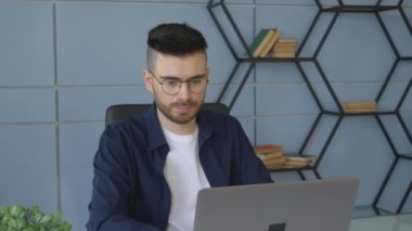 Gözlüklü genç bir erkek bilgisayar üzerinde çevrimiçi bir proje üzerinde çalışıyor, bir erkek programcı kod yazıyor ve parlak bir odada bir iç işlerinde yöneticilik yapıyor.