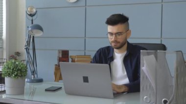 Yakışıklı beyaz adam, şık, rahat bir oturma odasında koltukta otururken dizüstü bilgisayarda çalışıyor. Evden çalışan Freelancer. İnternet taranıyor, Sosyal ağlar kullanılıyor