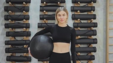 Siyah bir spor topuyla spor bir kızın portresi. Siyah bir top ve yerde duran taytlar, kameraya bakan parlak bir spor salonu.