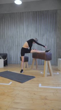 Jimnastik salonundaki jimnastik barlarında egzersiz yapan dikey video sporcusu genç bir kadın. Esneme egzersizleri, normal spor, siyah bir bluz ve tayt.