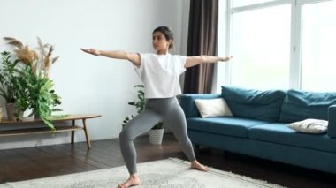 Genç Hintli kadın evinde yoga yapıyor. Pindasana pozuyla dizlerini göğsüne eğiyor. Tek ayak üzerinde durup denge egzersizi yapıyor.
