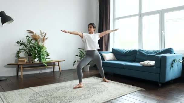 若いインド人女性がヨガをしていて 片方の脚に立ってバランスのための運動をしています 明るい部屋に家にいて 体を伸ばす — ストック動画