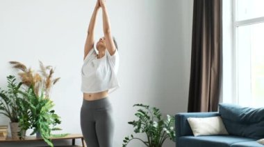 Genç atletik Hintli esmer kadın yoga yapıyor, kas ve eklemlerini esnetiyor, pencerenin yanında duruyor, evinde parlak bir odada.