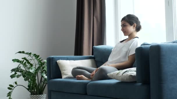 若いアスレチックインドのブルネットヨガの練習をしている女性 離れて見て息を飲んで 家の明るい部屋でソファに座って — ストック動画
