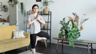 Genç bir Hintli kadın sabah evde parlak bir oda için meditasyon yapıyor. Tek bacaklı olmak, yoga yapmak, sabahları evde güneşli bir odada egzersiz yapmak, mutfağın arka planına karşı spor üniforması giymek,