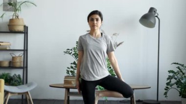 Spor kıyafetleri, pantolonlar ve bluzlar giyen, oturma odasında spor egzersizleri yapan, sağlık hizmeti veren ve ev kavramında zihinsel terapi uygulayan çok ırklı genç ve çekici bir kadın.