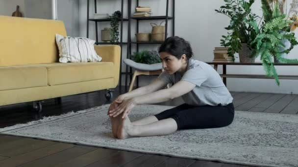 一位年轻的亚洲女人坐在地板上热身 伸展肌肉 锻炼身体 带着耳机呆在舒适的房间里 — 图库视频影像