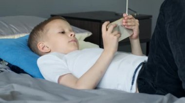 Bir çocuk telefon ekranına bakar, akıllı bir telefondan oyun oynar, parlak bir odada, yatakta sırtüstü uzanır.