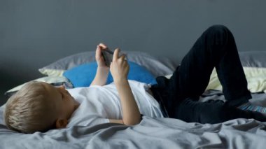 Çocuk akıllı telefonda oyun oynuyor, çocuk yatakta akıllı telefon oynuyor, çocuk telefon kullanıyor ve oyun oynuyor, bağımlı oyun oynuyor ve çizgi film oynuyor.