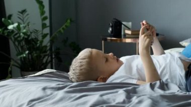 Yakından bakıldığında bir çocuk telefon ekranına bakar, akıllı bir telefonla oyun oynar, parlak bir odada, yatakta sırtüstü uzanır.