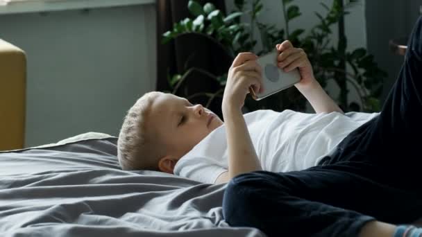 子供の男の子の肖像画携帯電話でゲームをプレイ画面を見て 明るい居心地の良い部屋で自宅でベッドの上に彼の背中に横たわる — ストック動画