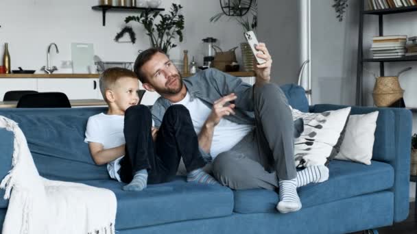 若い家族の父と子供が明るい部屋に座って ソファに座って スマートフォンを使って 子供は画面を見ています — ストック動画