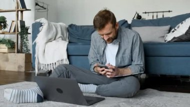 Genç bir erkek serbest çalışan, akıllı telefon kullanır, bir mesaj basar, evde çalışan bir serbest yazar, dizüstü bilgisayar kullanır, monitör ekranına bakar, yeni bir proje geliştirir.