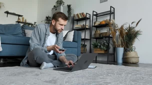 若いフリーランスの男は自宅で働き ノートパソコンを使用して モニター画面を見て 新しいプロジェクトを開発し 彼の手にスマートフォンを持っています — ストック動画