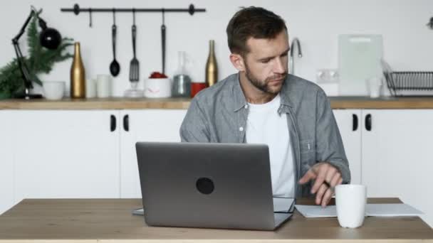 若い男が家のテーブルに座ってノートパソコンで作業するメモを書きます — ストック動画