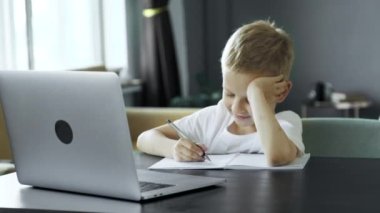 Portrait of adorable kid sitting alone doing homework with happy face, Cute child boy using pencil writing on white paper. Students doing spelling test at home. Homeschooling concept