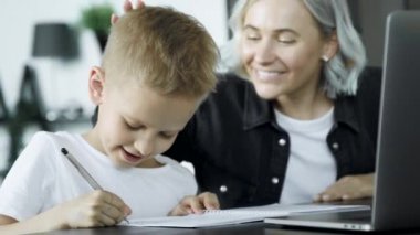 Close Up Mother Helping Son With Homework Sitting At Desk In Bedroom. The Child Does Homework and Uses A Computer, Is Engaged In Training, Sits at A Table in A Bright Cozy Room