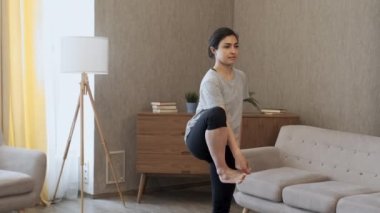 Bir Kadın Denge İçin Egzersiz Yapıyor, Yoga pozunda tek ayak üzerinde duruyor, Spor üniforması giyiyor, Evinde, rahat bir odada.