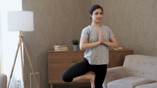 若いインド人女性が家の明るい部屋で瞑想しています 1本足で立ちヨガをしたり 明るい部屋で朝瞑想運動をする家の中で背景を背景に — ストック動画