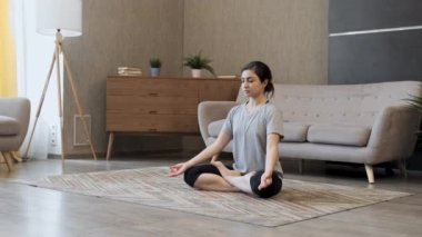 Spor giyimli atlet kadın oturma odasında yoga yapıyor. Nilüfer pozisyonunda meditasyon yapan esnek Hintli yetişkin gözleri spor minderinin üzerinde açık sağlıklı yaşam tarzının tadını çıkarıyor.