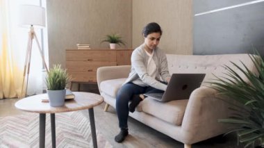 Gülümseyen Hintli kız evdeki kanepede dizüstü bilgisayar kullanıyor.