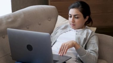 Kanepede dizüstü bilgisayar kullanan, düşünceli yüzlü, parlak bir odada koltukta uzanan Hintli kadını kapat.