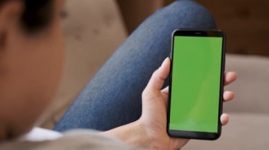 Kanepede oturan ve yeşil ekranlı akıllı telefon kullanan genç bir kadın.