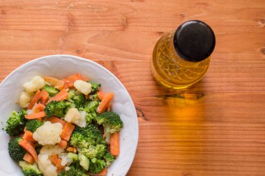 Karnabaharlı beyaz tabak, brokoli, havuç ve bir şişe yağ, ahşap zemin üzerinde izole edilmiş. Yakından görülüyor