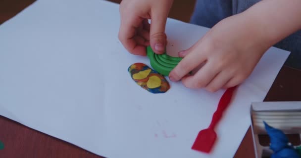 Um menino brinca com argila brilhante ou plasticina, sentado à mesa. — Vídeo de Stock