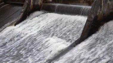 Geniş bir görüş, suyun köpüklü bir şelalede barajın üzerinden aktığını gösteriyor. Soldan sağa doğru panoramik bir manzara, dinamik enerjiyle akıyor..