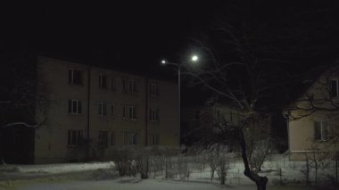 Karla kaplı zemin, çıplak ağaçlar ve karanlık pencereleri olan sakin bir kasabada sakin bir kış gecesi. Tek bir sokak lambasıyla hafifçe aydınlatılıyor. Sıcak ışık ve uzun gölgeler bırakıyor..