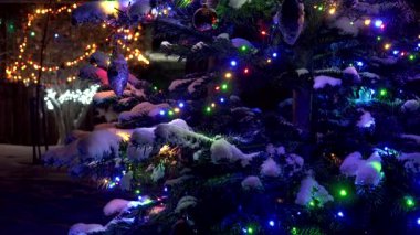 Geceleri karla kaplı bir Noel ağacı dalına yakın çekim. Renkli LED ışıklar, aydınlatıcı süsler ve taze kar. Arkaplanı çok renkli bokeh doldurur. Büyüleyici bir şenlik havası.